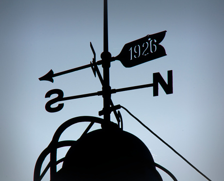 An English weathervane pointing at the sun, but silhouetted against approaching storm clouds, implying danger or trouble ahead.