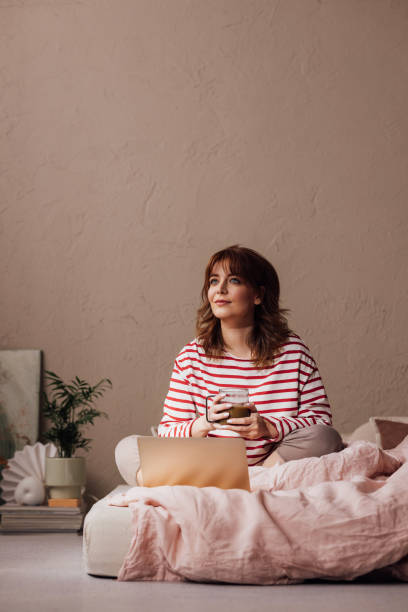 Morning Routine: Beautiful Ginger Woman Enjoying a Cup of Coffee in her Bed, while Using her Laptop
