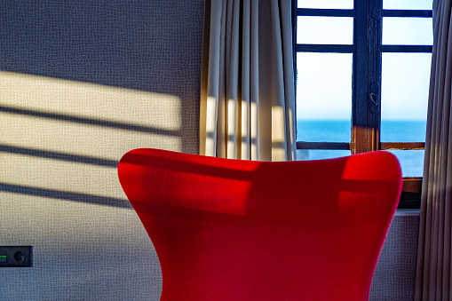 Asturias, Spain. Red armchair backrest next to a sunny window facing the sea. Minimalist, relaxed and summery scene.
