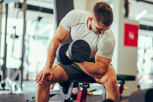 A strong sportsman is sitting in a sports center and doing dumbbell biceps curls while looking at his biceps. A muscular sportsman is sitting in a gym and flexing muscles during his training.