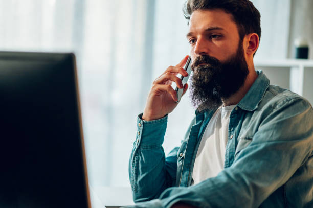 istock A bearded casual entrepreneur is sitting in his home office, looking at his computer and talking on the phone with an important client. 1492091353