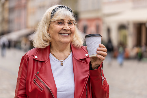Senior old woman enjoying morning coffee hot drink and smiling outdoors. Relaxing taking a break. Elderly grandmother walking in urban city center street drinking coffee to go. Town lifestyles outside