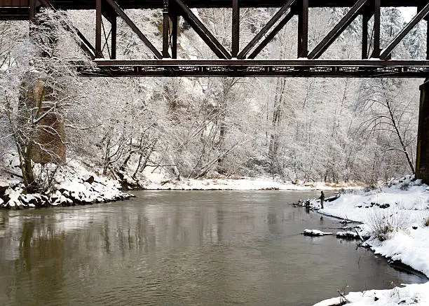 A lone fisherman in the distance fishes for salmon from the riverbank under a railroad trestle.  The scene is covered with fresh snow.