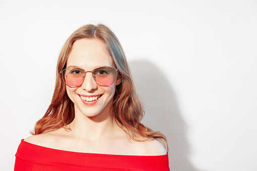 Close-up studio portrait of a happy young white woman with long brown hair in a red dress, wearing pink sunglasses, against a white background