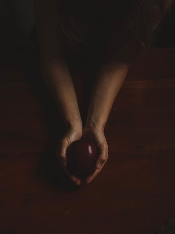 In the dimly lit room, a soft glow cascades upon a wooden table, revealing delicate, feminine hands gracefully clasping a luscious, crimson apple. As the overhead perspective captures the scene, every contour and texture of the hands and fruit come alive, painting a picture of mystery and allure. The contrasting elements of darkness and vibrant red evoke a sense of anticipation, inviting contemplation of the apple's enticing secrets hidden within.