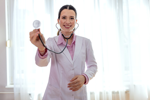 A young Caucasian female doctor is looking at the camera with a smile and holding up a stethoscope.