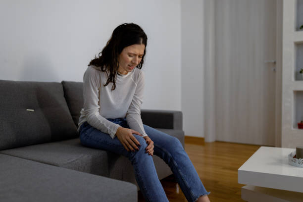 I hit my knee on the edge of the table A young Caucasian woman with painful face expression sitting on a living room couch and holding hands on her knee erythema nodosum stock pictures, royalty-free photos & images
