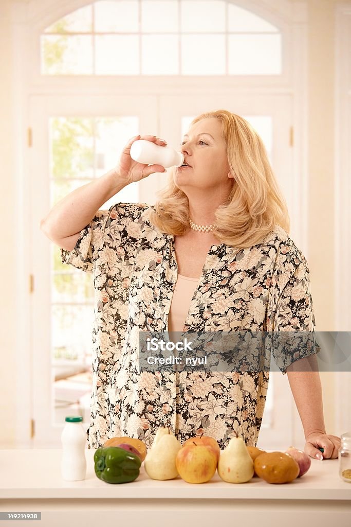 Woman drinking yogurt in kitchen Senior woman standing in kitchen, drinking yogurt in bright background, with vegetables and fruits on counter... 50-59 Years Stock Photo