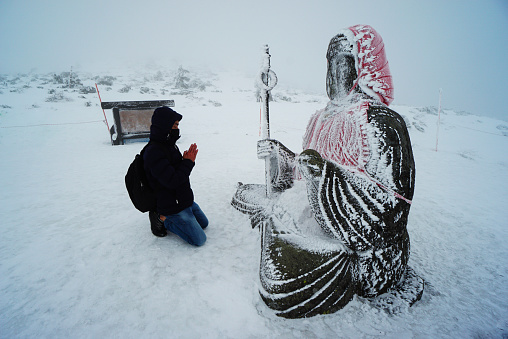 Asian man praying to Buddha statue on the mountain peak during blizzard