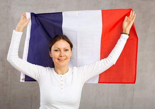 Happy smiling middle-aged woman waving national flag of France while looking at camera at gray wall background