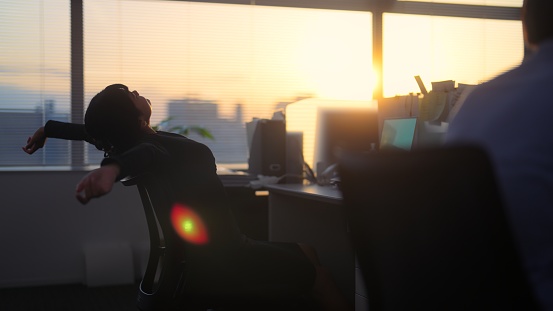 A businesswoman is taking a break from working and stretching her arms and relaxing in an office during sunset.