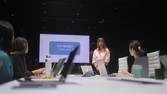 A businesswoman is using graphs and visuals on monitor and giving presentation in front of colleagues in a modern office.