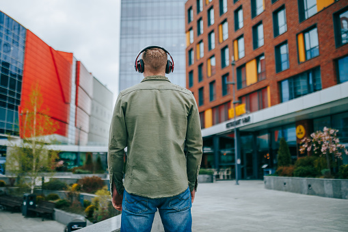 A good-looking hispanic guy listening to music while taking a walk in the city, captured from behind with the building background. Rear view of man enjoying music while walking in the city