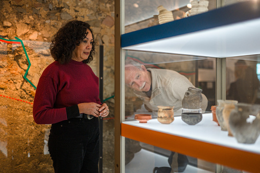 Mature multiracial female historian and her senior male coworker, both in casual clothes, deciding on the details of a Roman earthenware exhibition. 3/4 length.