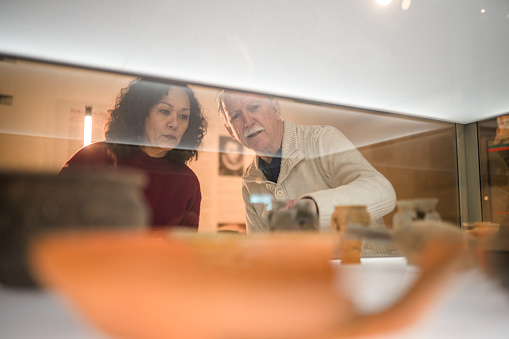 Close up of a handsome senior man and a pretty,  mature woman looking at the exhibition of Roman earthenware in blurred foreground. Looking away, shot through glass.