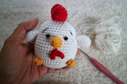 Hand holding crochet handmade of tiny white chicken doll on fluffy background with needle and yarn behind it.
