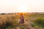 Stylish woman with straw hat dancing at oat field in sunset light. Atmospheric happy moment. Young female in rustic linen dress relaxing in evening summer countryside, rural life