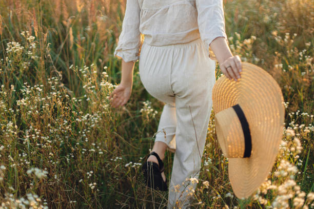 Stylish boho woman with straw hat in hand close up among wildflowers in sunset light. Atmospheric moment. Summer delight and travel. Young female in rustic linen cloth walking in summer meadow stock photo
