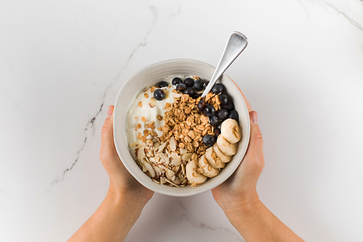 A woman holds a bowl of homemade granola, Greek yogurt, blueberries, almonds and a banana. Healthy breakfast concept, recipe.