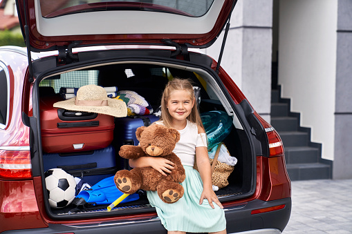 Portrait of little girl sitting in car trunk full of luggage