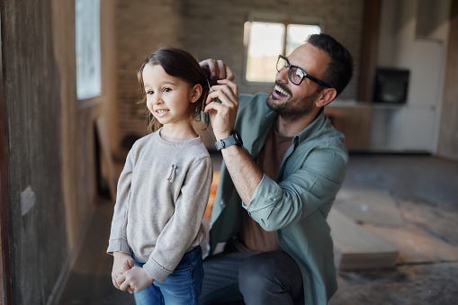 Happy single father making a ponytail for his small daughter during home renovation process. Focus is on girl.