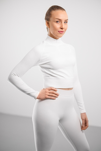 Portrait of smiling young woman in athleisure standing with hand on hip against white background