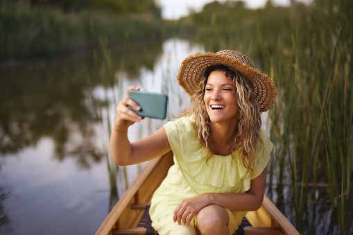 Happy woman taking a selfie with cell phone while spending a summer day in canoe at lake.