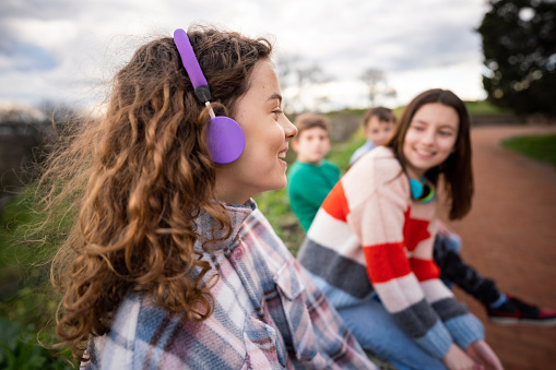 Group of happy teenage boys and girls talking while sitting in park. Focus is on happy girl whit headphones