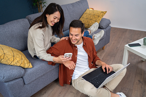 Young Caucasian man working on a laptop from home, while his girlfriend of Japanese ethnicity using a mobile phone while relaxing on the sofa
