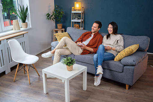 Young multiracial couple, Japanese woman and Caucasian man, sitting on the sofa in the living room and watching TV
