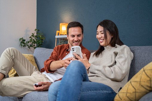 The young multiracial couple, relaxing on the sofa in their modern apartment, while watching interesting content on mobile phone