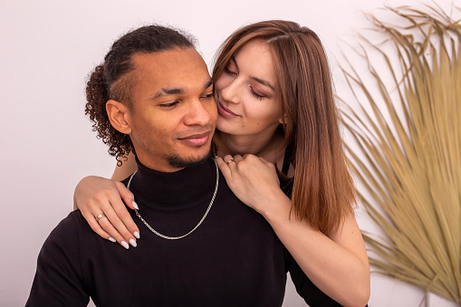 Portrait of smiling beautiful multiracial couple, in black clothes, are sitting near the wall in the room. Close up. Copy space