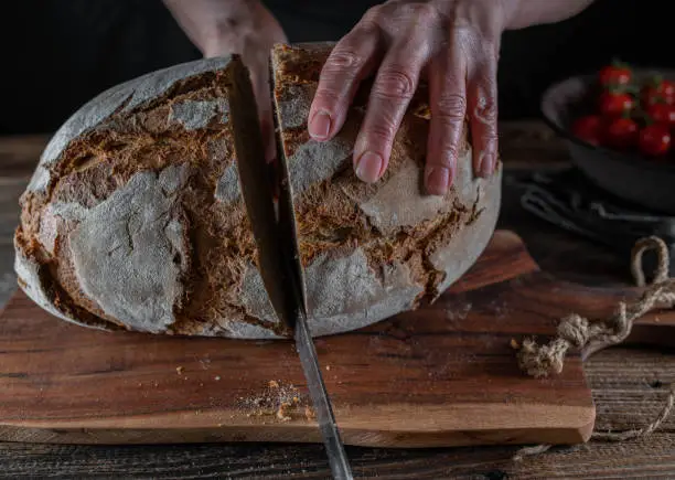 Rustic german sourdough bread is cutting in half by womans hands on a old fashioned cutting board on wooden tablen. Closeup, front view