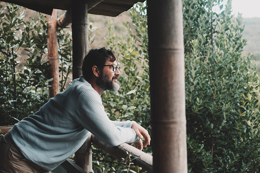 Serene adult man looking the nature outside home in terrace patio having relax and connection with nature. One house owner living in recycled material wood house. Handsome male with glasses