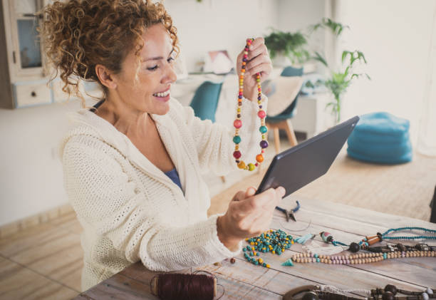 Happy woman indoor leisure hobby work activity producing beads accessories necklaces showing her jewel in video call using tablet and wireless connection technology. New business at home lifestyle stock photo