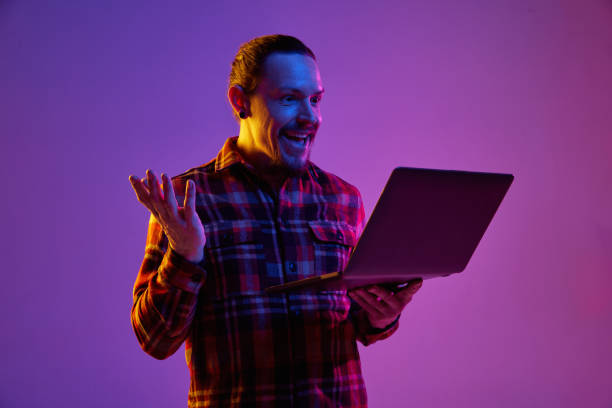 Portrait of happy smiling man using laptop for work or communication against purple studio background in neon light