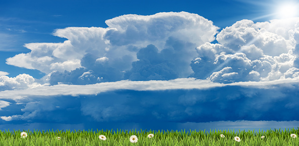 Beautiful cumulus clouds (cumulonimbus) on blue sky with torrential rain and sunbeams over a green meadow with daisies flower.