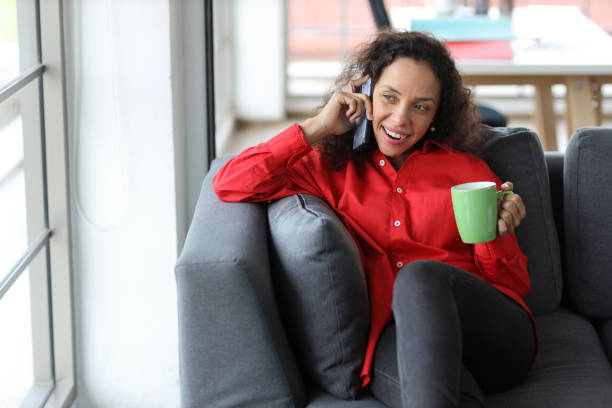 istock Smart and active latin woman sitting on sofa and using internet on computer with smart mobile phone while drinking coffee in living room. Lifestyle with technology 1492238404