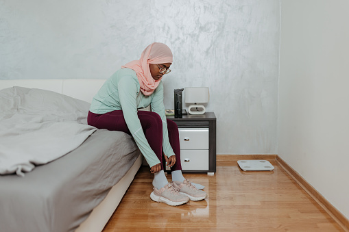 Witness the harmonious pursuit of fitness and faith as a young Muslim woman, donning a hijab, finds balance in her life. In this serene scene, she starts her exercise regimen by sitting on her bed and stretching