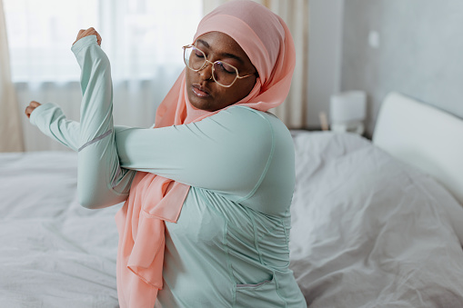 Discover a moment of tranquility as a young Muslim woman, donning a hijab, starts her day by sitting on her bed and stretching her arms