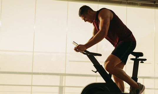 Young man cycling on a stationary bike in a gym, smashing his cardio goals with focus and dedication. Male athlete pushing himself to achieve optimal health, fitness and wellbeing.