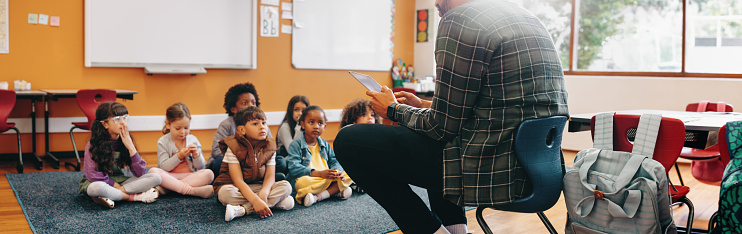 Man teaching an elementary school class using a digital tablet. Teacher is sitting in front of his students and is reading a story from an ebook. Education and child development in a multiethnic school.