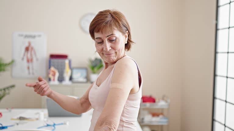 Mature hispanic woman doing thumbs up with band aid on arm at clinic