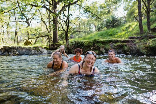 Group of women wild swimming in the Lake District, North East of England. They are in a river, enjoying time outdoors. They are laughing together in the cold water.