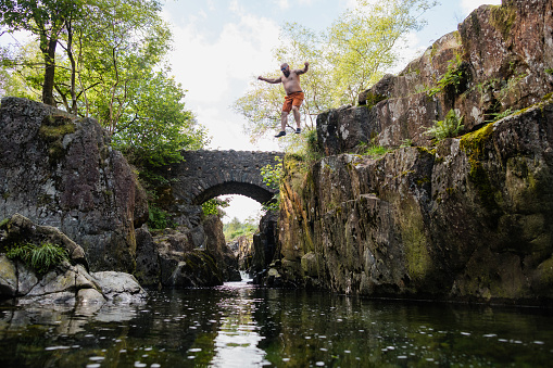 Man wild swimming in the Lake District, North East of England. He is jumping into a river from a high river bank, enjoying time outdoors.