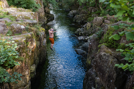 Wide high angle view of a man wild swimming in the Lake District, North East of England. He is jumping into a river from a high river bank, enjoying time outdoors.