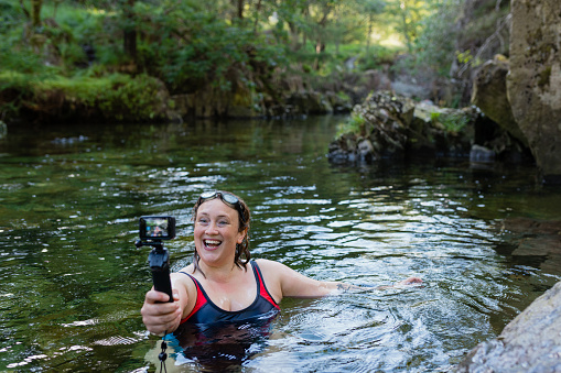 Woman wild swimming in the Lake District, North East of England. She is in a river, enjoying time outdoors, using a go pro to vlog her time there.