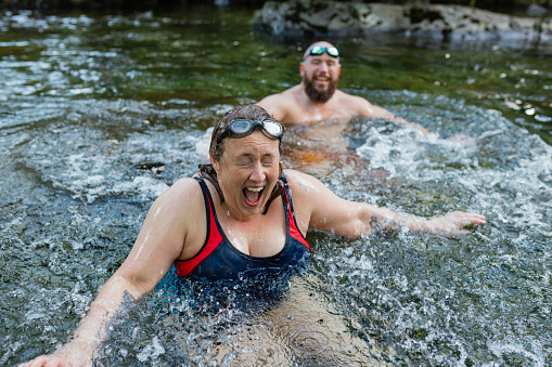 Couple wild swimming in the Lake District, North East of England. They are in a river, enjoying time outdoors. The woman is laughing with her eyes closed and mouth open.
