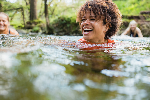Group of women wild swimming in the Lake District, North East of England. They are in a river, enjoying time outdoors. The focus is on one woman who is laughing.