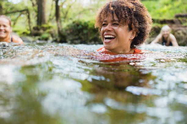 Summer Soak Group of women wild swimming in the Lake District, North West of England. They are in a river, enjoying time outdoors. The focus is on one woman who is laughing. woman cold water swimming stock pictures, royalty-free photos & images